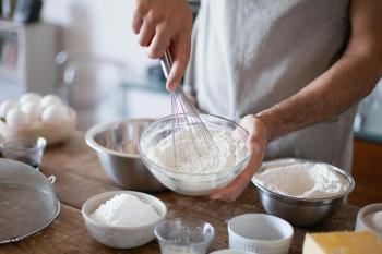 A baker is whisking a bowl of flour.  Other bowls of ingredients are on the table in front of them.