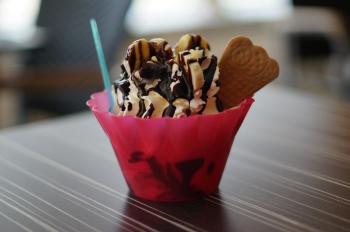 Vanilla ice cream with bananas and chocolate syrup drizzled on top.  A triangle shaped cookie is sticking out of the side.  The ice cream is in a bright pink bowl with a blue spoon.
