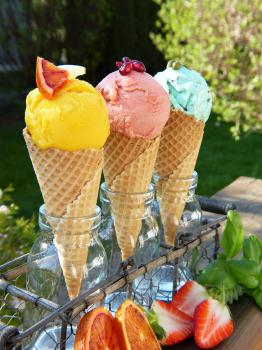 Three ice cream cones sitting in a display case above fresh strawberries.  The cones are waffle cones, and the ice cream is three different bright colors.  Yellow, pink, and blue.