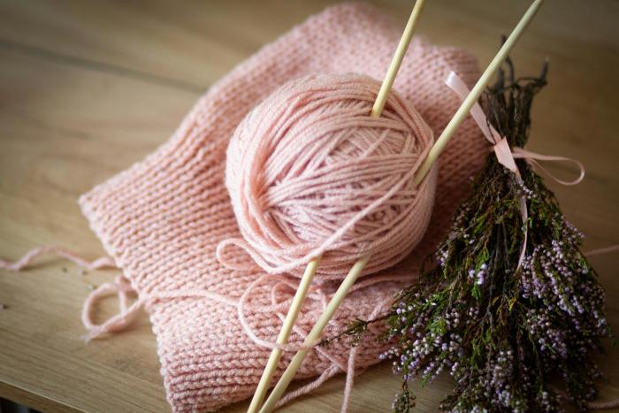 Pink ball of yarn, knitted piece of fabric in the same color, wooden needles, and a bunch of lavender tied with a pink ribbon.