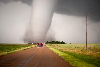 A massive tornado that hit near Dodge City, KS in 2016, at the end of a long dirt road.  Three cars are stopped on the road in front of the twister.  The funnel cloud is wider than the road itself, and you can see the dust and debris whirling around the base.